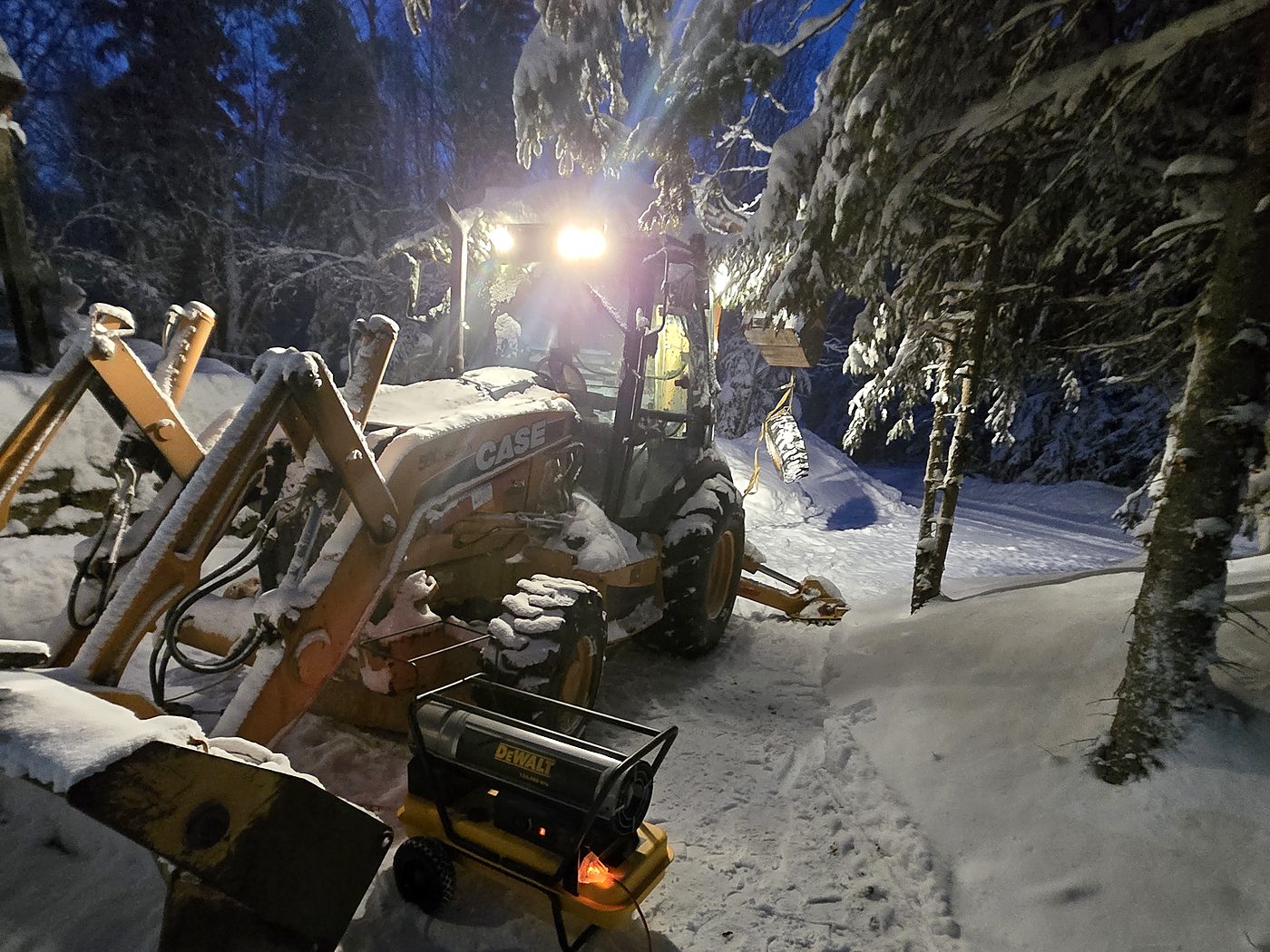 Snow-covered Case backhoe loader at dusk with its work lights on, parked on a snowy trail between spruce trees, a DeWalt heater on the snow in front of it