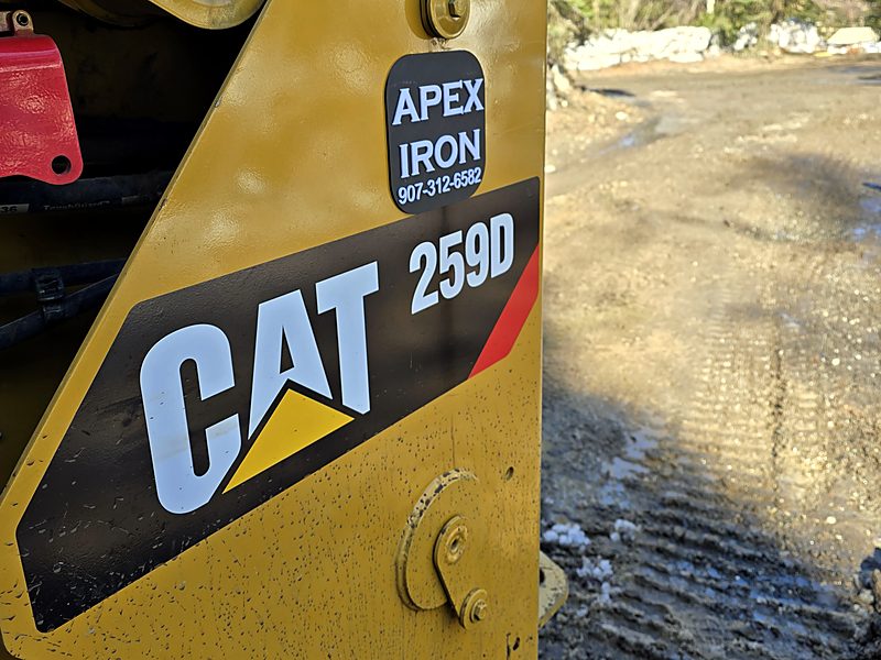 Close-up of the side of a yellow Cat 259D compact track loader wearing an Apex Iron decal with the phone number 907-312-6582